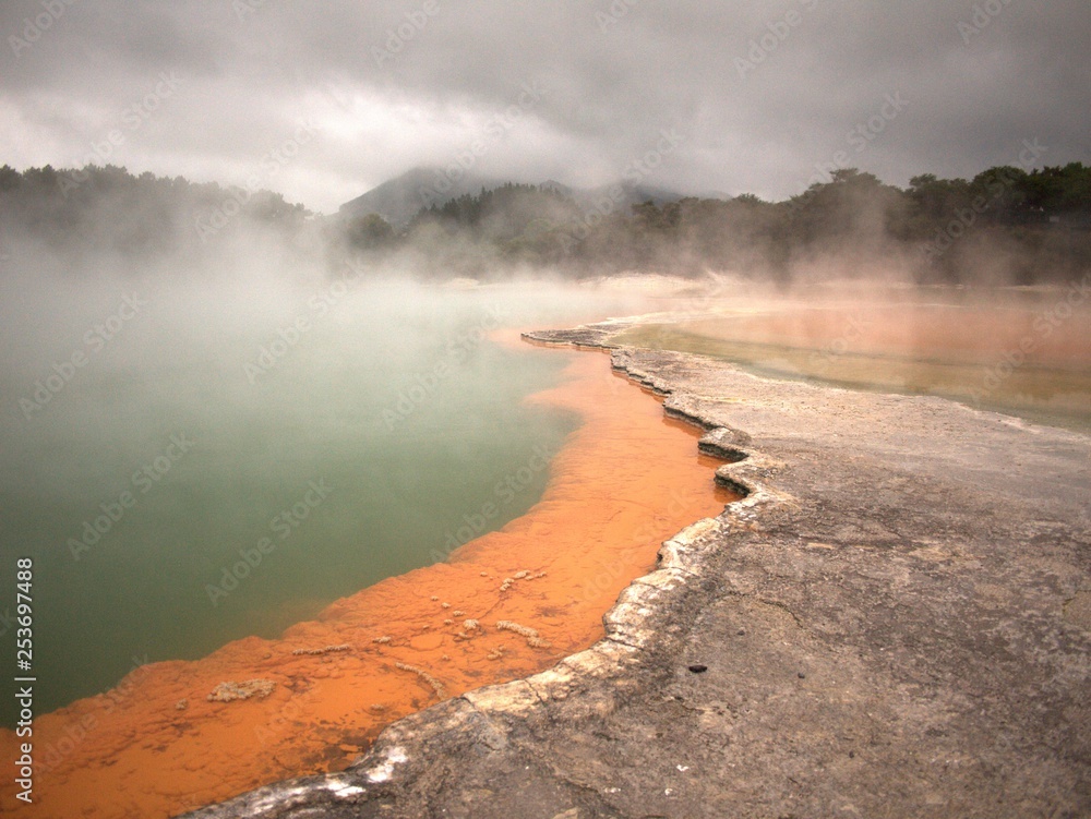 Volcanic rock pool with layers of gray, green, and orange rock with steam rising from water and trees and hills in background under grey, cloudy sky
