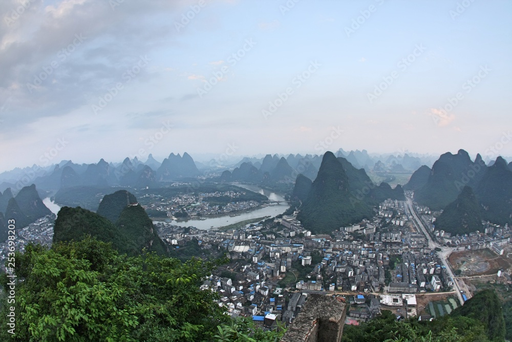 Limestone karst peaks in Yangshuo, Guangxi, China with town low-rise and high-rise buildings in valley and river underneath blue sky with hazy smog in air