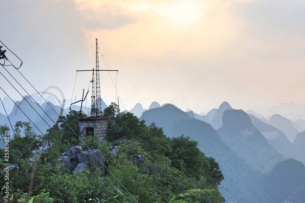 Metal, steel electrical tower for television broadcast with cables in front of karst peaks in Yangshou, China with orange setting sun in cloudy grey sky and smog in air