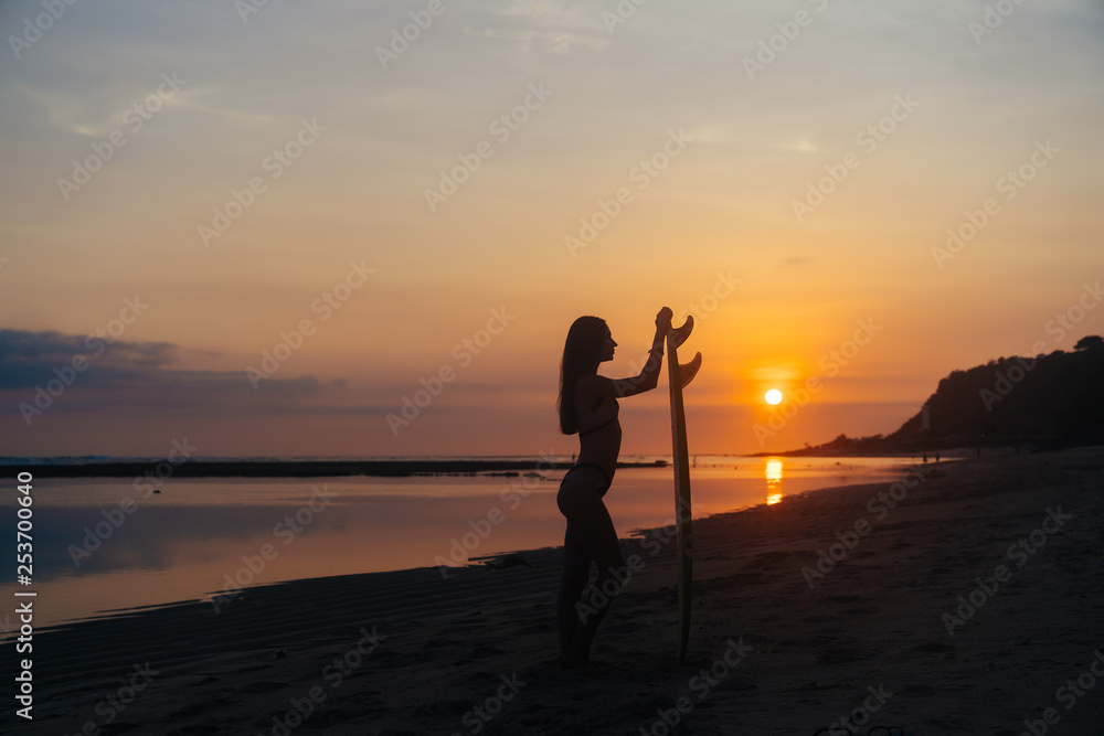 Silhouette of slim girl with surfboard in hands at beach on background of beautiful sunset