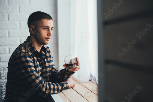 A young guy with a glass of whiskey leaned on the windowsill looking into the glass photo