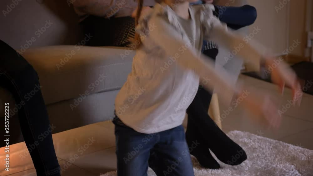 Close-up portrait of small caucasian girl with braids watching movie attentively and pointing finger on it with her mother and sister on background. Close-up portrait of small caucasian girl with braids watching movie attentively and pointing finger on it with her mother and sister on background.