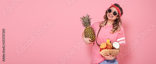 Beautiful young girl in pink t-shirt and glasses, holds a full straw bag of fruit on pink background