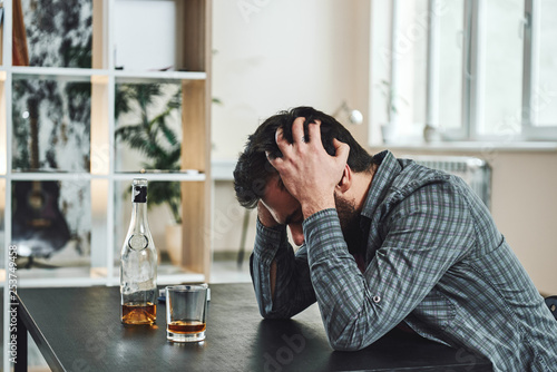 The most common way people give up their power is by thinking they don’t have any. Drunk man with glass and bottle of whiskey sitting at table in kitchen. Drinking alone. Male alcoholism concept photo