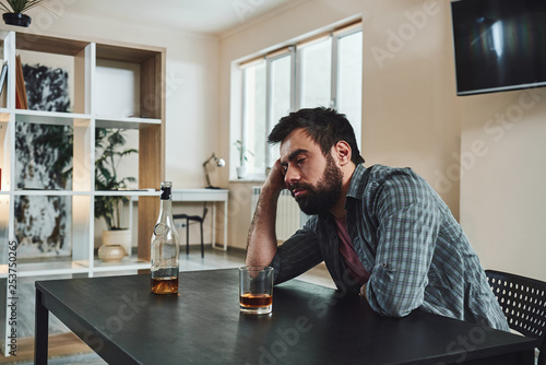 Alcohol does not solve problems but becomes a problem in itself. Drunk man with glass and bottle of whiskey sitting at table in kitchen. Drinking alone. Male alcoholism concept