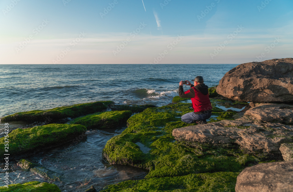 Traveler on a rocky seashore overgrown with green algae taking pictures on smartphone