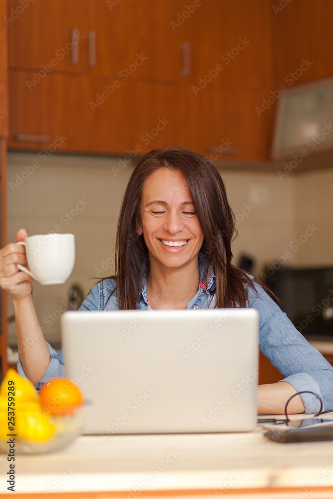Smiling mid adult woman drinking coffee and using laptop in kitchen Smiling mid adult woman drinking coffee and using laptop in kitchen
