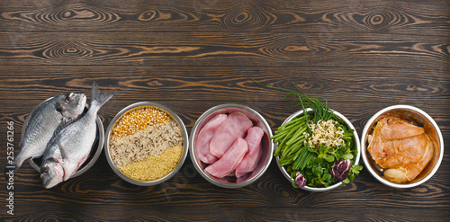 Panorama banner with healthy pet food ingredients with chopped raw turkey, fish, groats, greens and grains in individual bowls on brown wooden background. Flat lay. photo
