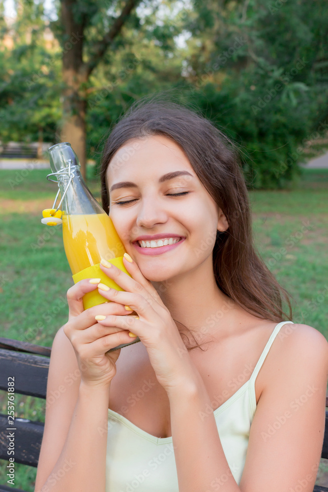 Smiling girl is refreshing with a cold bottle of lemonade near the forehead in hot day in the park Smiling girl is refreshing with a cold bottle of lemonade near the forehead in hot day in the park