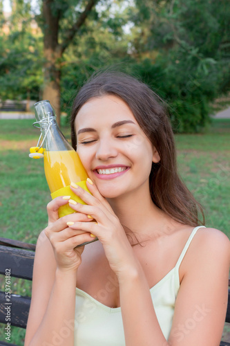 Smiling girl is refreshing with a cold bottle of lemonade near the forehead in hot day in the park