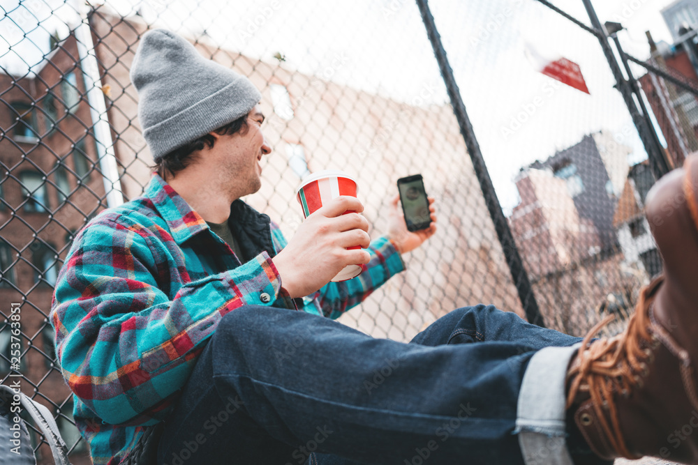 Naklejka premium Young trendy man sitting on the street sidewalk resting with cup of hot coffee and using smartphone for video link. Casual man calling on video link his friends