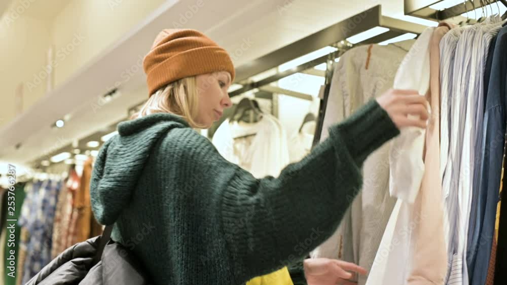 A girl in a green sweater and a yellow hat walks through a store of things and chooses what to buy. Touches things on hangers and looks at price tags A girl in a green sweater and a yellow hat walks through a store of things and chooses what to buy. Touches things on hangers and looks at price tags