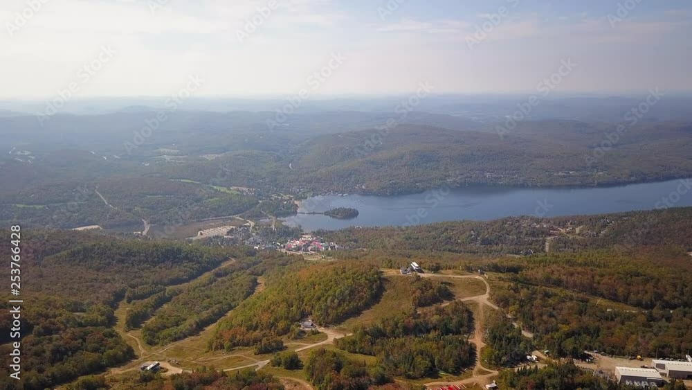 Smooth aerial / drone shot moving forwards over the forest on a hike path during summer season with green, yellow and orange trees, a lake, a small town and mountains on the background. Smooth aerial / drone shot moving forwards over the forest on a hike path during summer season with green, yellow and orange trees, a lake, a small town and mountains on the background.