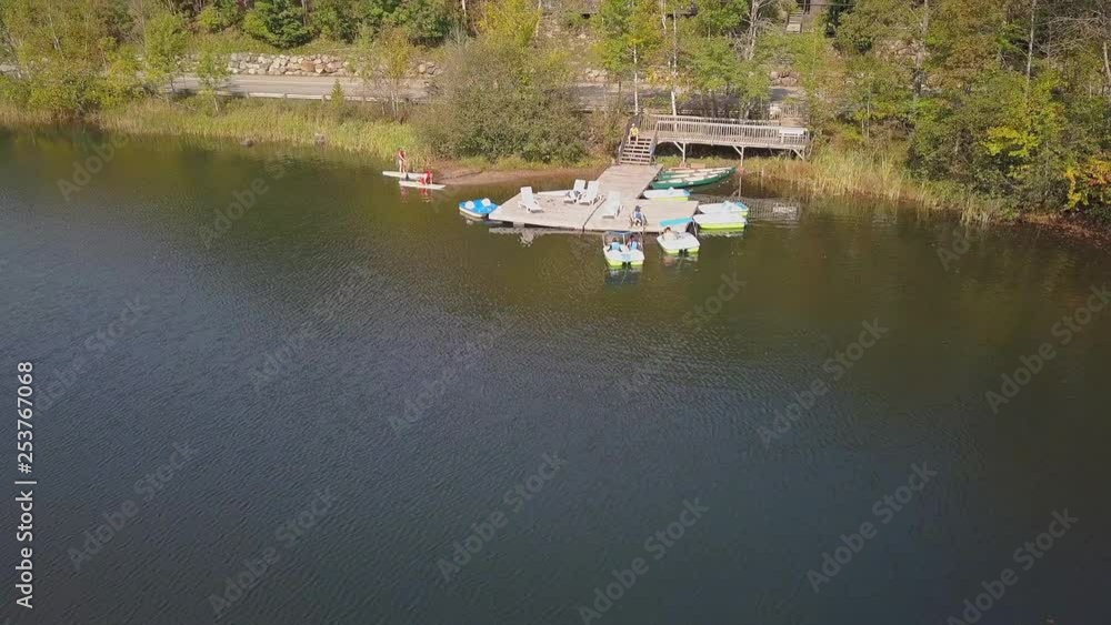 Pedal boats and stand up pads on the lake near a dock with canoes during summer season with green, yellow and orange trees on the background. Pedal boats and stand up pads on the lake near a dock with canoes during summer season with green, yellow and orange trees on the background.