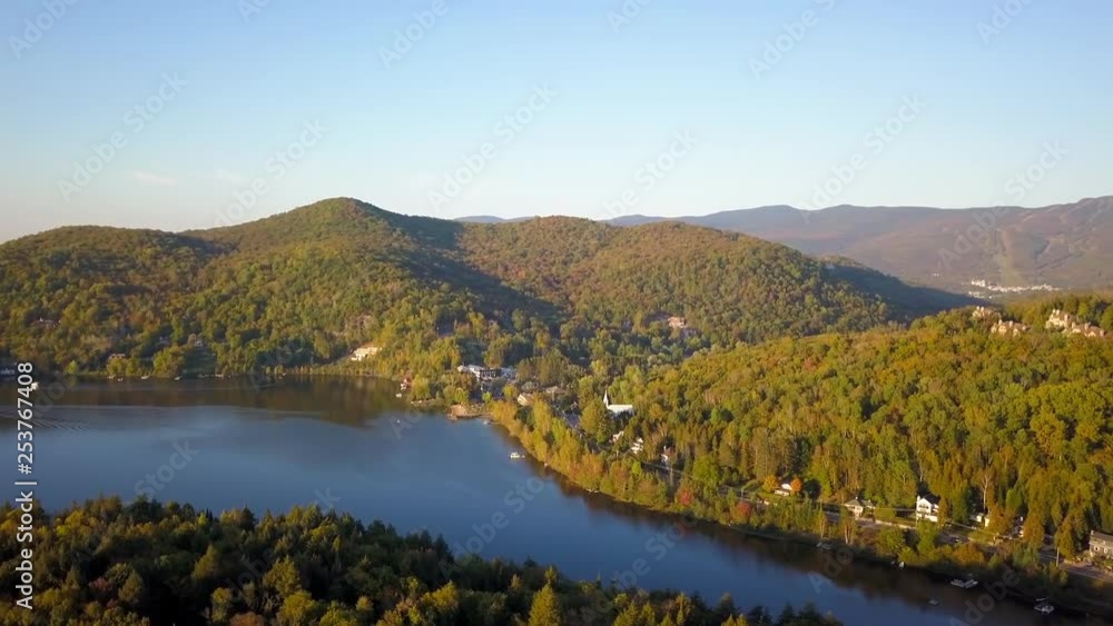 Smooth panning aerial / drone shot over the forest on a driveway, cottages and a lake during summer season with green, yellow and orange trees on the background.