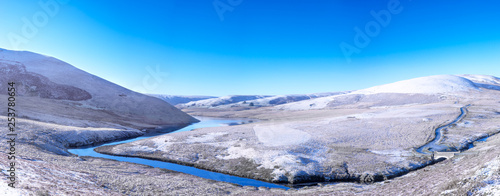 Elan Valley in Wales showing Afon Elan river flowing through a snowy panorama of a winter scene of mountains and blue sky.