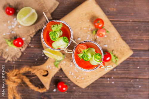 Two glasses of fresh organic tomato juice decorated with raw tomatoes, cucumber and leaves on a rustic wooden cutting board