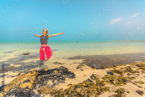 Happy blonde woman jumping at turquoise clear sea at Persian Gulf near Al Areesh Fishermens villages, Northerm of Qatar.Traveler blonde tourist visits abandoned village, Middle East, Arabian Peninsula photo
