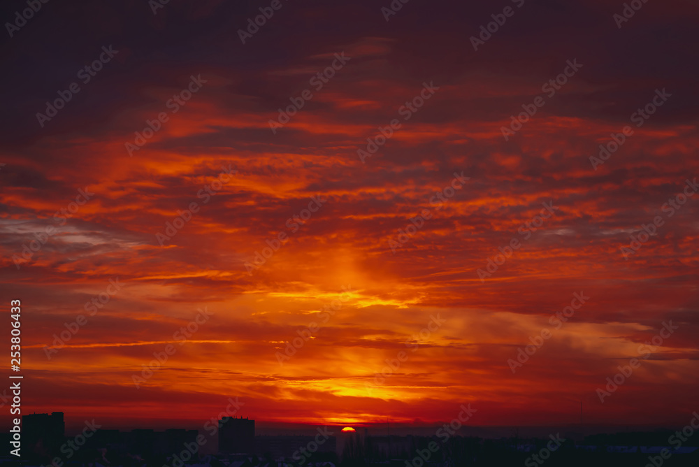 Cityscape with vivid fiery dawn. Amazing warm dramatic cloudy sky above dark silhouettes of city buildings. Orange sunlight. Atmospheric background of sunrise in overcast weather. Copy space.