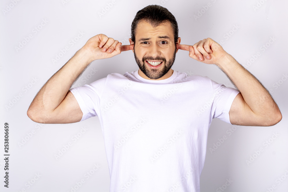 Handsome man in white t-shirt over white background, smiling, covering his ears with hands Handsome man in white t-shirt over white background, smiling, covering his ears with hands
