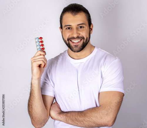 A handsome man in a white T-shirt on a white background, smiling, holding pills in his hand