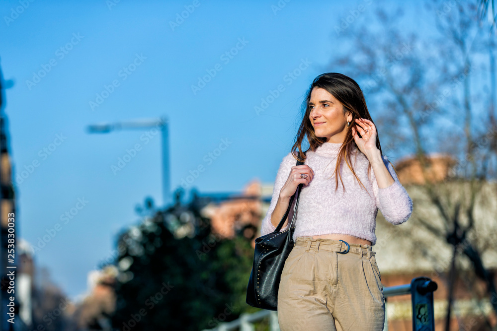 Front view of smiling beautiful young woman touching hair and standing in the street while looking away in a sunny day Front view of smiling beautiful young woman touching hair and standing in the street while looking away in a sunny day