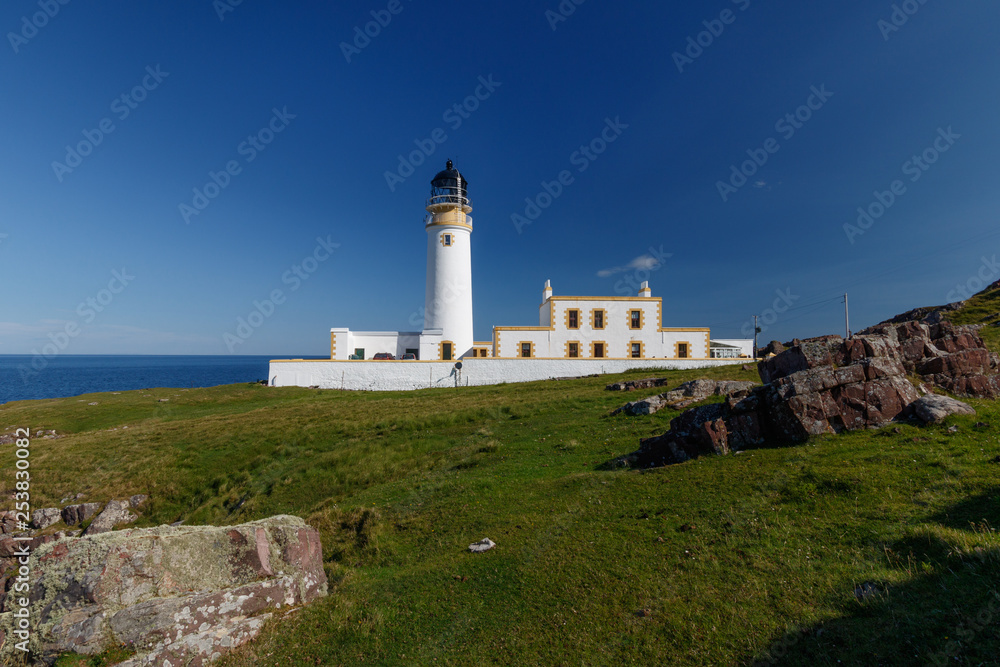 Rua Reidh Lighthouse with keepers house and walls on grassy hillside along the coast. Rocks in the foreground and blue sky. Rua Reidh Lighthouse with keepers house and walls on grassy hillside along the coast. Rocks in the foreground and blue sky.