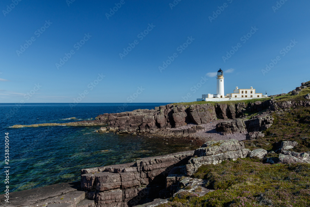 Rua Reidh Lighthouse with keepers house on a rocky coastline with blue sky.