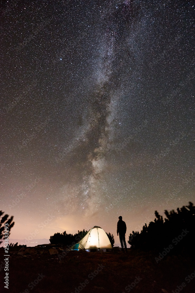 Silhouette of male hiker having a rest at summer night camping on the top of the mountain beside illuminated tourist tent. Happy man traveller enjoying view of night sky full of stars and Milky way.