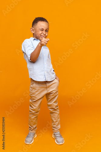 Quadro su tela Full length image of confident cool dark skinned schoolboy in fashionable wear standing at yellow studio wall, pointing fore finger at camera, winking at you
