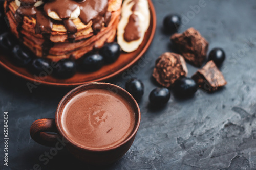 Hot chocolate drink in a cup and pancakes with banana, chocolate sauce and grapes in plate, on dark background