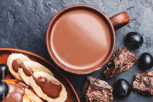 Hot chocolate drink in a cup and pancakes with banana, chocolate sauce and grapes in plate, on dark background, top view