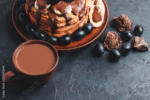 Hot chocolate drink in a cup and pancakes with banana, chocolate sauce and grapes in plate, on dark background