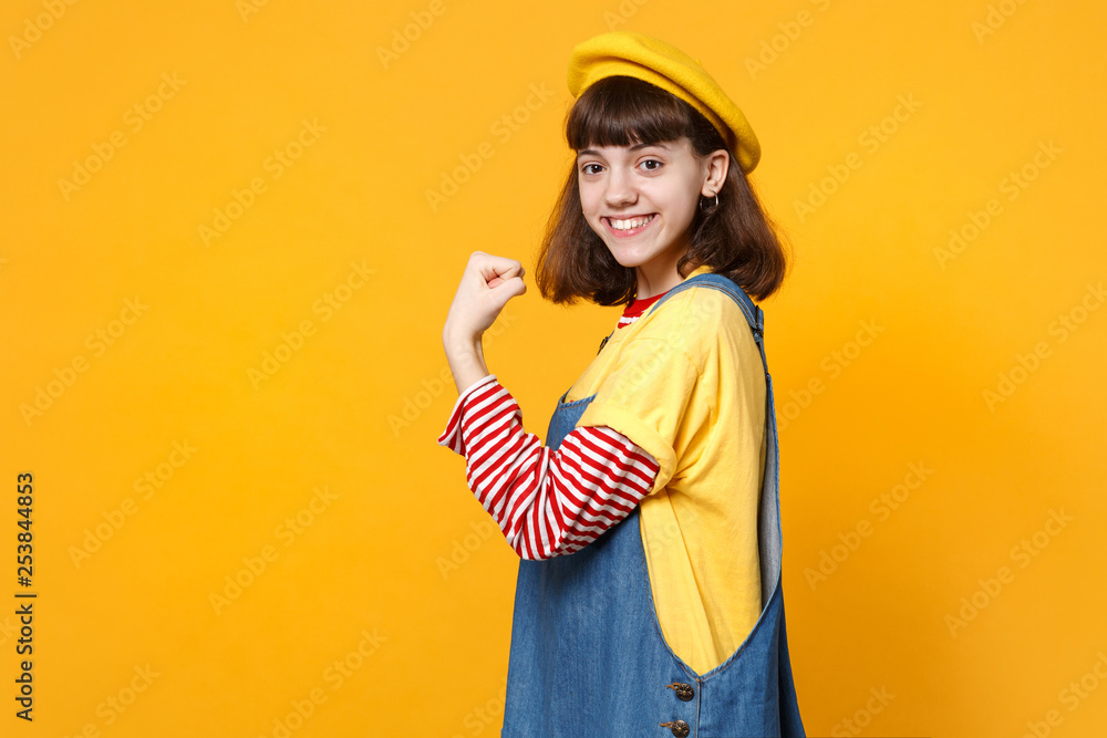 Portrait of beautiful girl teenager in french beret, denim sundress showing biceps, muscles isolated on yellow wall background in studio. People sincere emotions lifestyle concept. Mock up copy space. Portrait of beautiful girl teenager in french beret, denim sundress showing biceps, muscles isolated on yellow wall background in studio. People sincere emotions lifestyle concept. Mock up copy space.