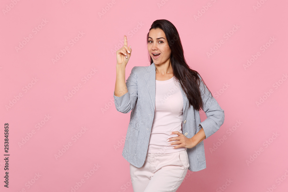Portrait of excited young woman in striped jacket holding index finger up with great new idea isolated on pink pastel wall background. People sincere emotions, lifestyle concept. Mock up copy space.