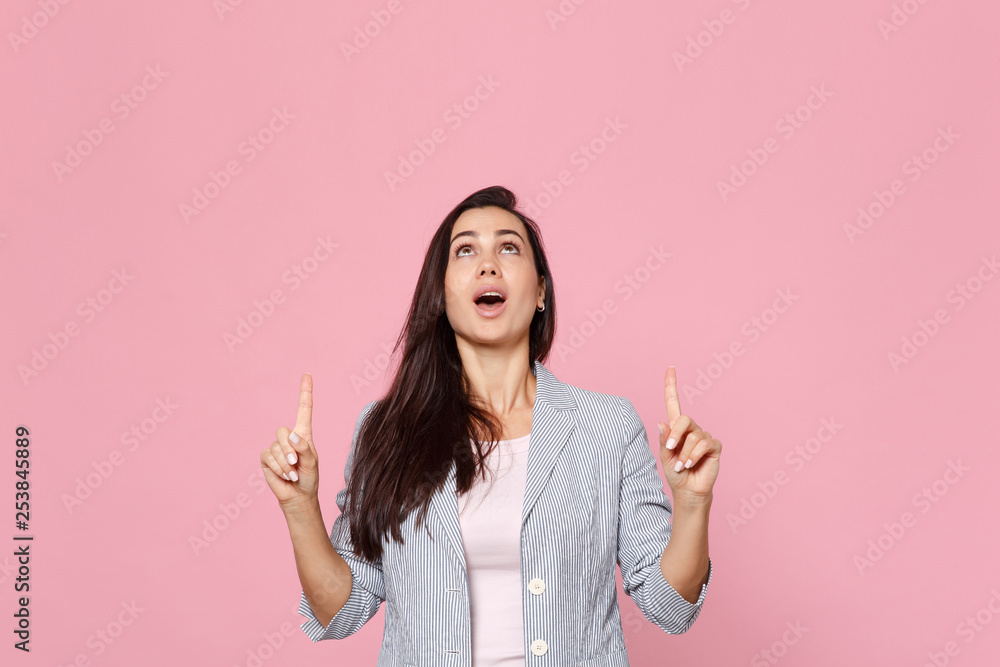 Portrait of amazed young woman in striped jacket looking, pointing index fingers up isolated on pink pastel wall background in studio. People sincere emotions, lifestyle concept. Mock up copy space. Portrait of amazed young woman in striped jacket looking, pointing index fingers up isolated on pink pastel wall background in studio. People sincere emotions, lifestyle concept. Mock up copy space.