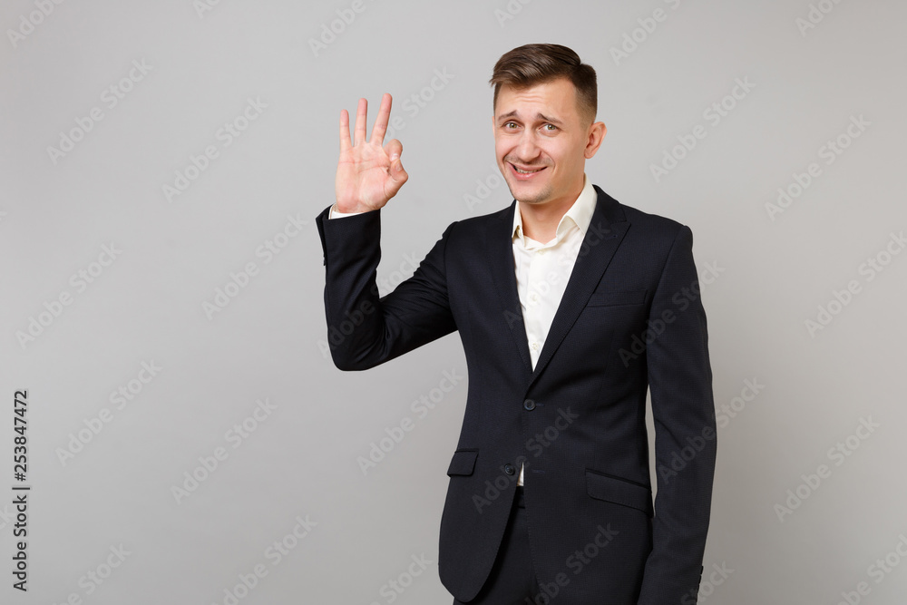 Portrait of satisfied young business man in classic black suit and shirt showing OK gesture isolated on grey wall background in studio. Achievement career wealth business concept. Mock up copy space.