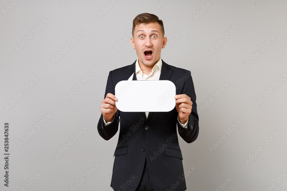 Shocked young business man in suit keeping mouth wide open, holding empty blank Say cloud speech bubble for promotional content isolated on grey background. Achievement career wealth business concept. Shocked young business man in suit keeping mouth wide open, holding empty blank Say cloud speech bubble for promotional content isolated on grey background. Achievement career wealth business concept.
