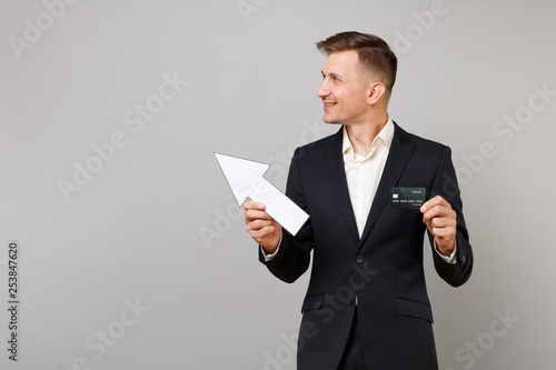 Young business man in classic black suit showing aside with arrow, looking aside, holding credit bank card isolated on grey background. Achievement career wealth business concept. Mock up copy space.