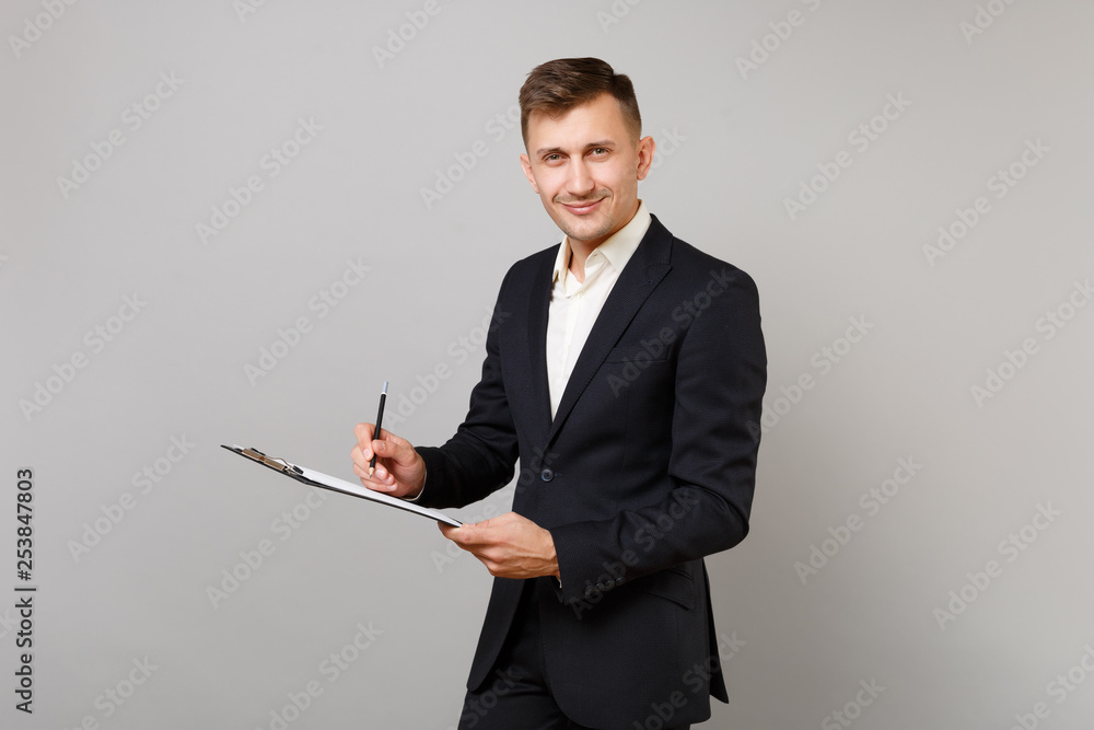 Successful young business man in classic black suit, shirt writing notes on clipboard with papers document isolated on grey background. Achievement career wealth business concept. Mock up copy space. Successful young business man in classic black suit, shirt writing notes on clipboard with papers document isolated on grey background. Achievement career wealth business concept. Mock up copy space.