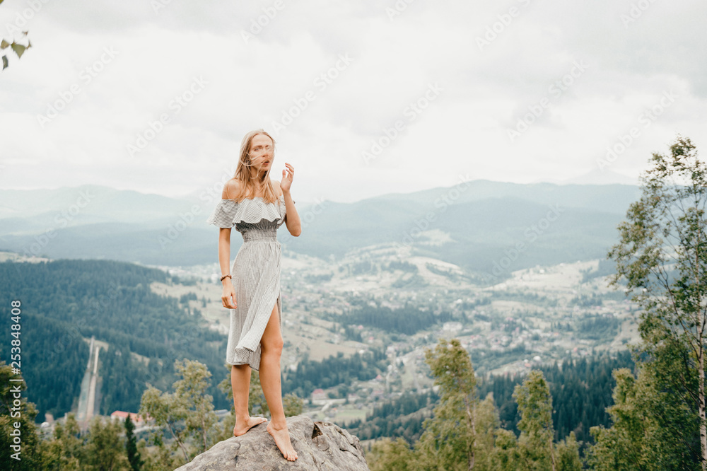 Naklejka premium Young beautiful barefoot blonde girl with long hair in summer dress standing on top of conquered mountain at stone and enjoying fabulous landscape scenic view with mountains and village in valley