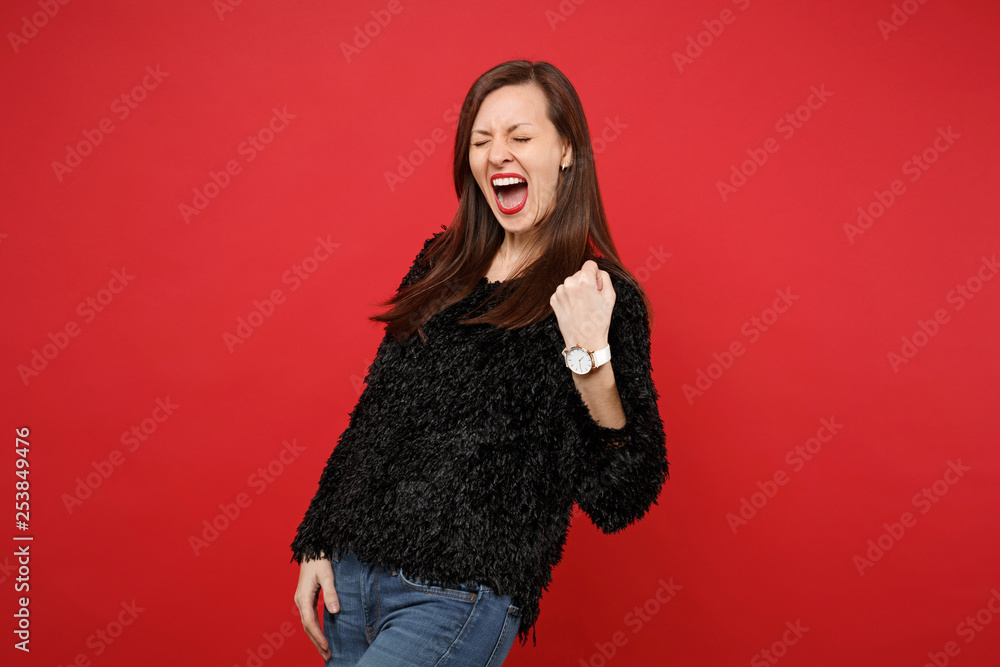 Overjoyed young woman in black fur sweater doing winner gesture, keeping eyes closed, screaming isolated on bright red wall background. People sincere emotions, lifestyle concept. Mock up copy space. Overjoyed young woman in black fur sweater doing winner gesture, keeping eyes closed, screaming isolated on bright red wall background. People sincere emotions, lifestyle concept. Mock up copy space.