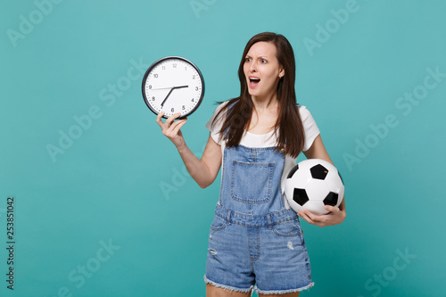 Irritated young woman football fan holding soccer ball, round clock isolated on blue turquoise wall background in studio. Time is running out. People emotions, sport family leisure lifestyle concept.