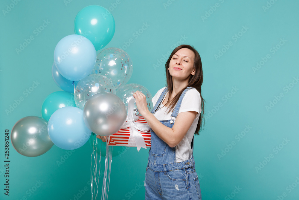Fototapeta premium Girl with closed eyes hold red striped present box with gift ribbon celebrating with colorful air balloons isolated on blue turquoise background. Valentine's Day Women's Day birthday, holiday concept.