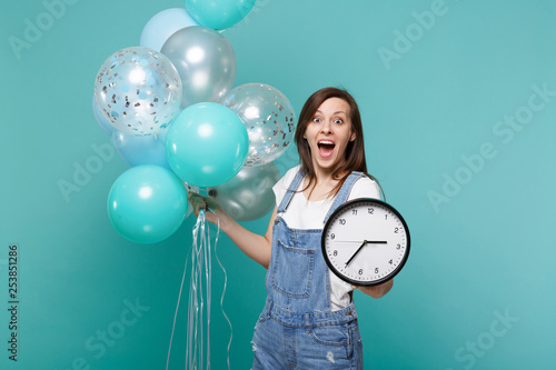 Surprised young woman keeping mouth wide open, holding round clock, celebrating with colorful air balloons isolated on blue turquoise wall background. Birthday holiday party, people emotions concept. photo