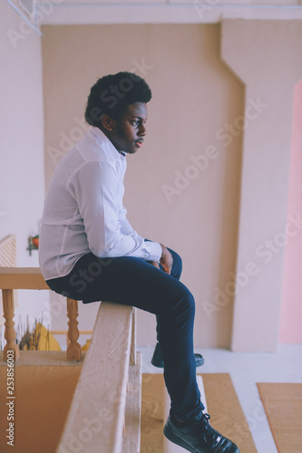 Lifestyle portrait of handsome young black man sitting at parapet at his decorated in african style room with plants near wall and stairs. Indoor portrait of happy dark-skinned nigerian male resting. © benevolente
