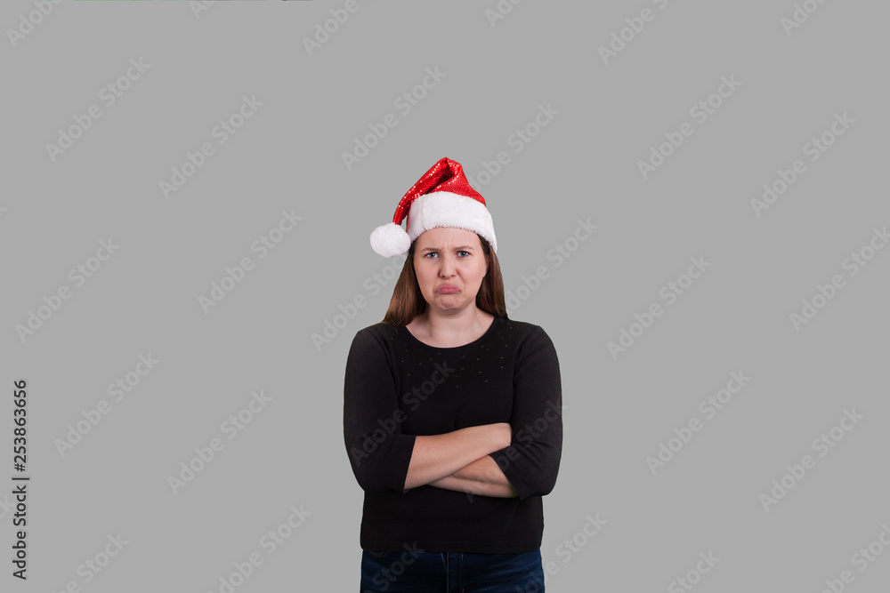 youthful women arms crossed pouty face wearing Santa Claus hat on solid gray background 
