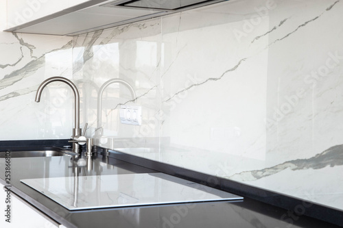 Close up of white glossy kitchen with black quartz countertop and marble tile backsplash. Build-in hidden incorporated hood and undermounted sink. 