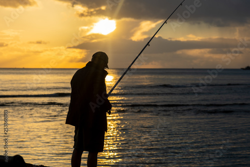 Fototapeta Naklejka Na Ścianę i Meble -  An unrecognizable man fishes while a beautiful sunset creates a stunning silhouette.