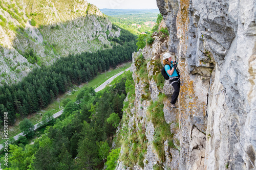 Female climber switching carabiners on a via ferrata route, vertical wall high above the valley, with a road behind. Klettersteig route at Baia de Fier, Pestera Muierilor, Romania, Gorj county. photo